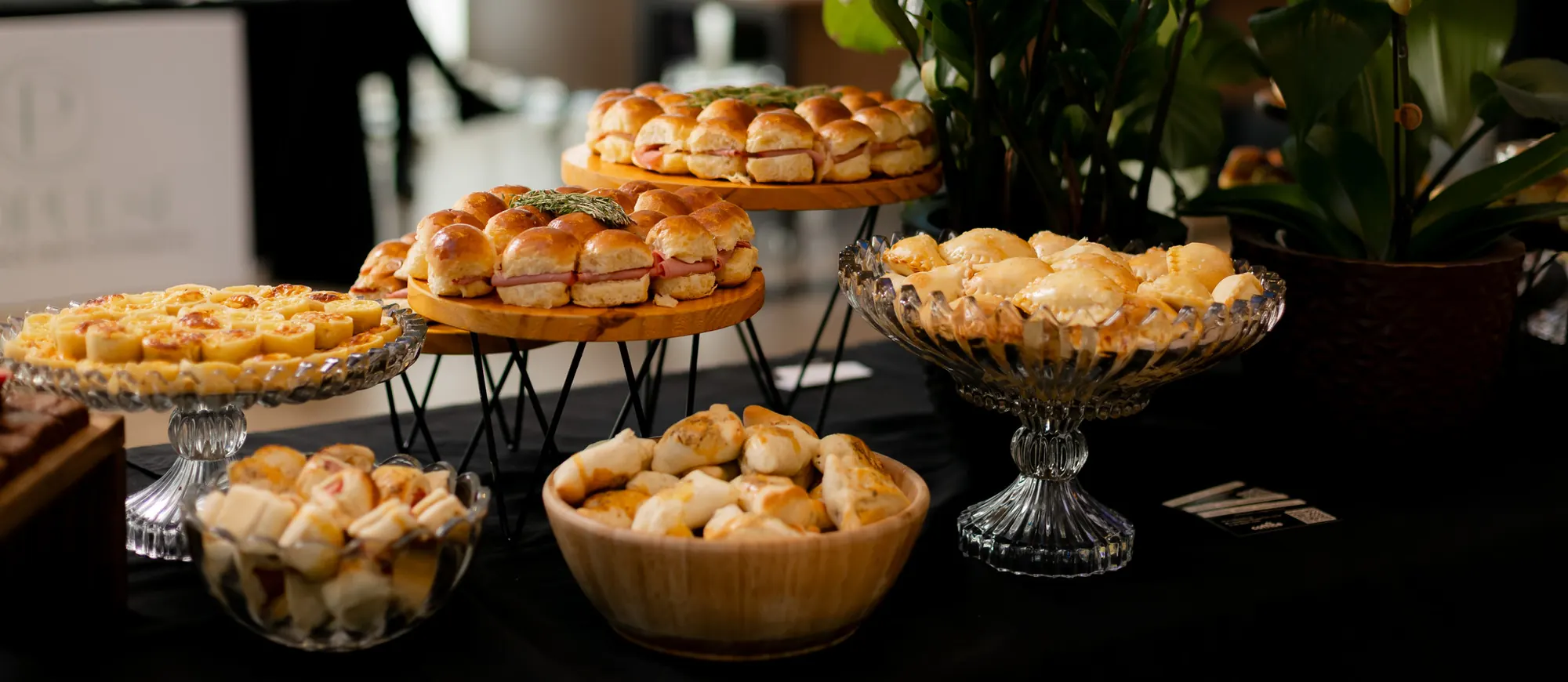 Mesa de coffee break montada pela Coffice num evento de empresa, com mini sanduíches, quiches e salgados em louças de vidro e madeira e uma orquídea