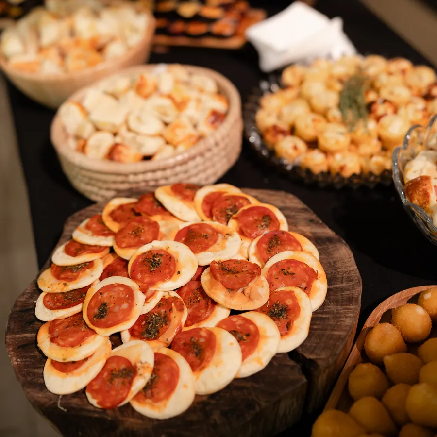 Mini pizzas de calabresa sobre uma tábua rústica, com outros salgados ao fundo